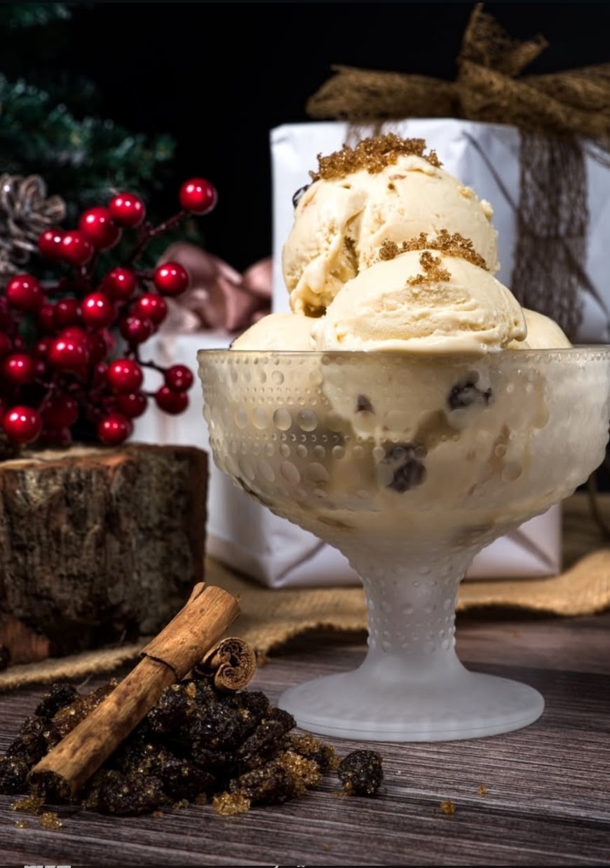 A glass bowl filled with scoops of vanilla ice cream topped with brown sugar and dark chocolate pieces, styled on a rustic wooden surface with cinnamon sticks, red berries, and wrapped gifts in the background. This photo was taken by our friends at Hovdenak Distillery.